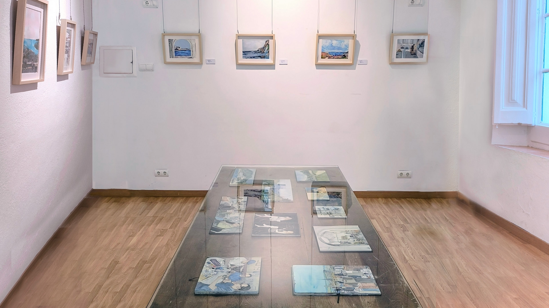 Vista de la sala petita de l’exposició “Moments”, amb obres de petit format a les parets i una taula de vidre amb llibretes d’esbossos com a inici del recorregut expositiu.
